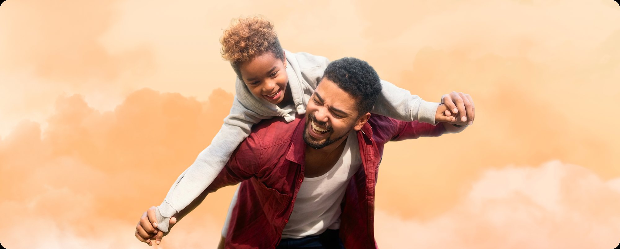 A father carrying his laughing daughter on his back, arms spread, against a warm sunset sky